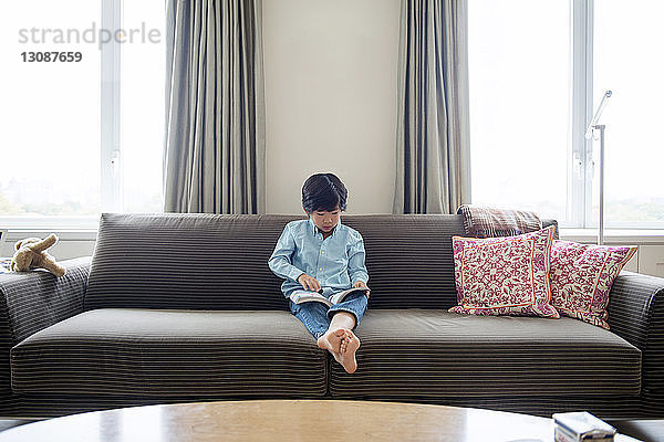 Junge liest Buch  während er zu Hause auf dem Sofa sitzt