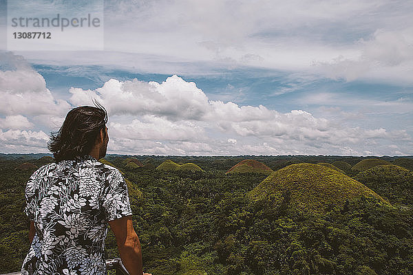 Rückansicht eines Mannes  der Chocolate Hills betrachtet  während er am Geländer vor bewölktem Himmel steht