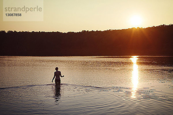 Rückansicht einer Frau im Bikini  die bei Sonnenuntergang im See steht