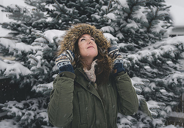 Frau schaut auf  während sie im Pelzmantel bei Schneefall an Bäumen klebt