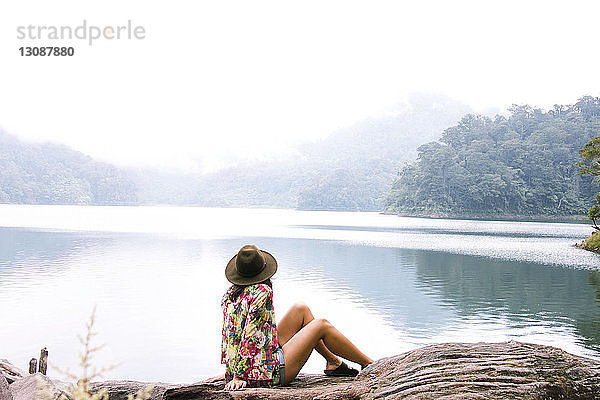 Seitenansicht einer Frau mit Hut  die bei nebligem Wetter auf einem Baumstamm am See sitzt