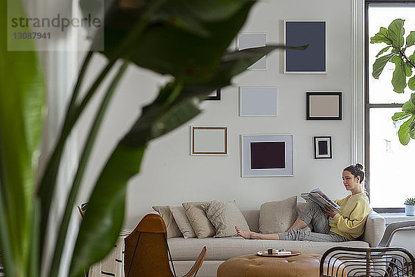 Seitenansicht einer Frau  die eine Zeitschrift liest  während sie zu Hause auf dem Sofa sitzt