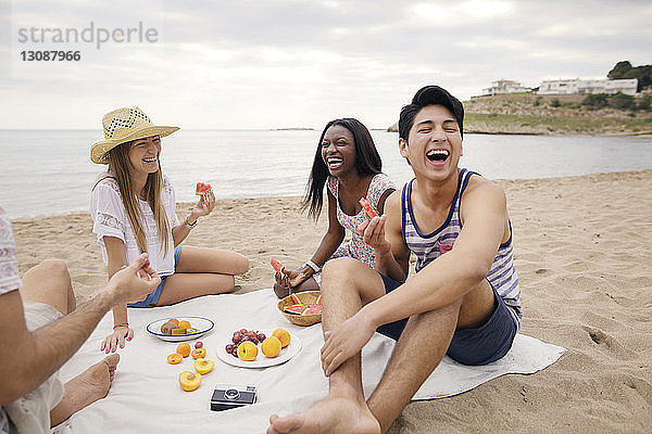 Fröhliche Freunde essen Früchte  während sie am Strand sitzen