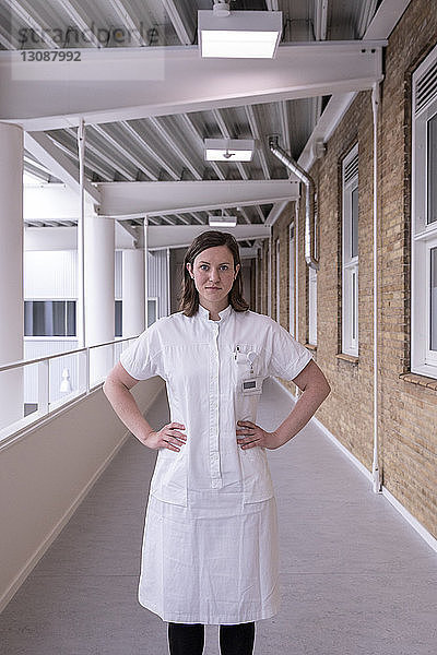 Porträt einer selbstbewussten Ärztin mit Händen auf der Hüfte im Krankenhauskorridor stehend