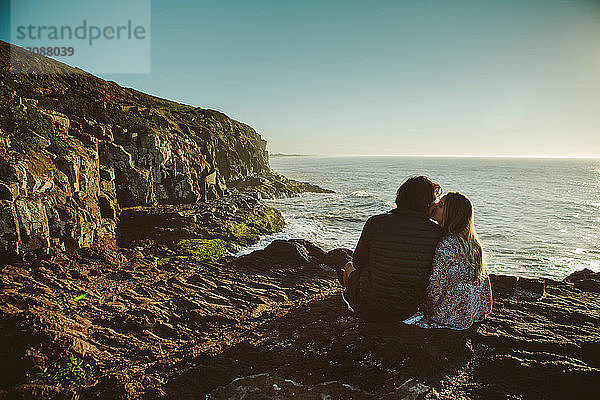 Rückansicht eines Paares  das sich küsst  während es auf einem Felsen am Meer vor klarem Himmel sitzt