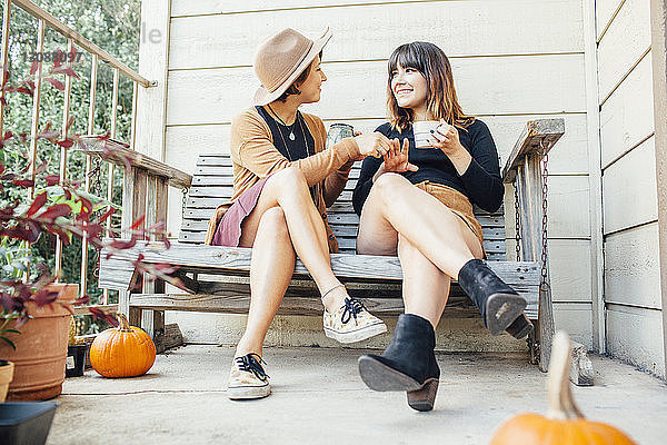 Freundinnen unterhalten sich in voller Länge  während sie auf einer Holzbank in der Veranda sitzen