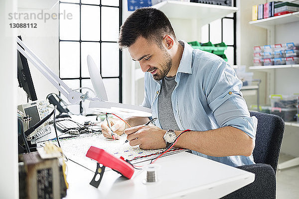 Lächelnder Techniker beim Löten von Leiterplatten am Tisch im Elektroniklabor