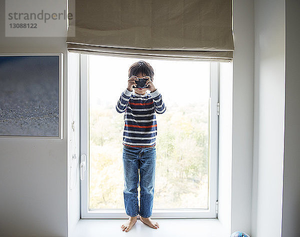 Fröhlicher Junge spielt zu Hause mit der Kamera auf dem Fensterbrett