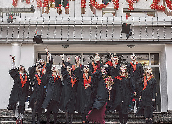 Niedriger Blickwinkel auf glückliche Studenten in Graduationskleidern  die auf Stufen am Gebäude stehen