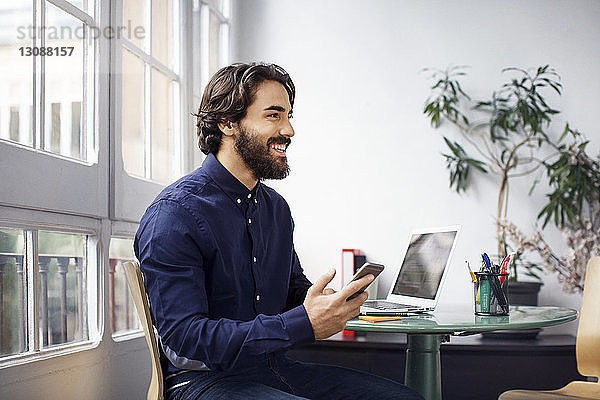 Nachdenklicher Geschäftsmann hält Smartphone in der Hand  während er im Büro auf einem Stuhl sitzt