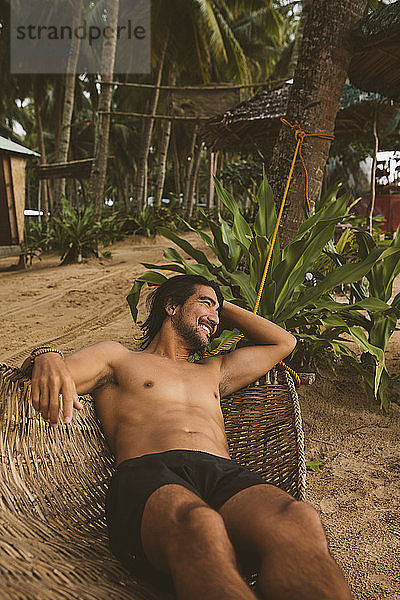 Hochwinkelansicht eines Mannes ohne Hemd  der beim Entspannen auf einer Hängematte am Strand wegschaut