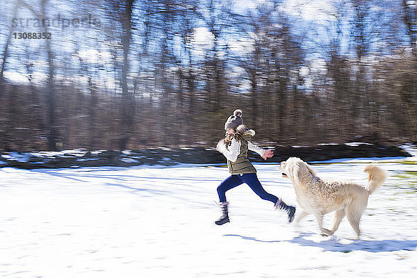 Seitenansicht eines Mädchens mit Hund  das auf einem schneebedeckten Feld gegen kahle Bäume läuft