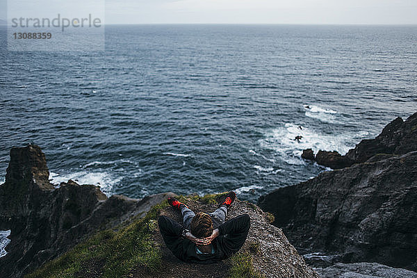 Hochwinkelansicht eines Wanderers  der mit den Händen hinter dem Kopf auf einer Klippe vor einer Meereslandschaft sitzt