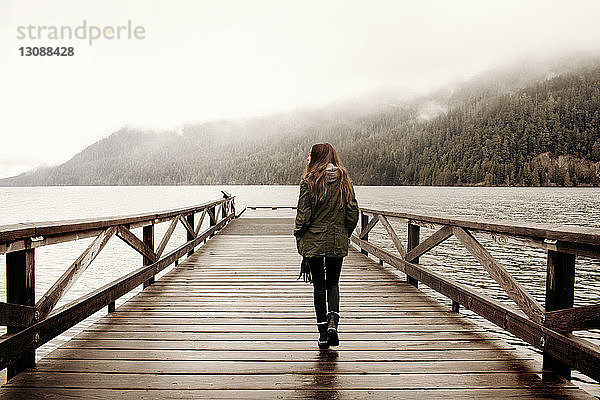 Rückansicht einer Frau  die bei nebligem Wetter auf einer Strandpromenade am See spazieren geht
