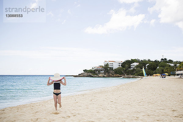 Mädchen hält Hut  während sie am Strand gegen Himmel und Meer läuft
