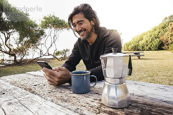 Glücklicher Mann mit Kaffee auf Holztisch  der mit seinem Handy gegen den Himmel sitzt