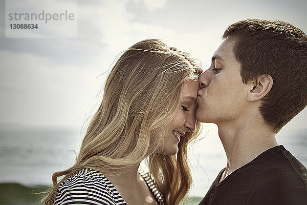 Seitenansicht eines am Strand stehenden Mannes  der die Stirn seiner Freundin küsst