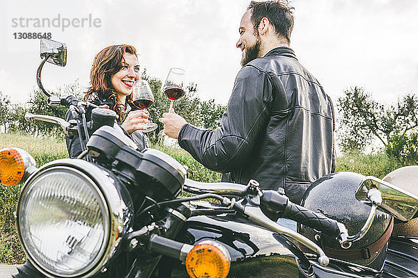 Glückliches Paar trinkt auf Weingläser  während sie mit dem Motorrad gegen den Himmel stehen