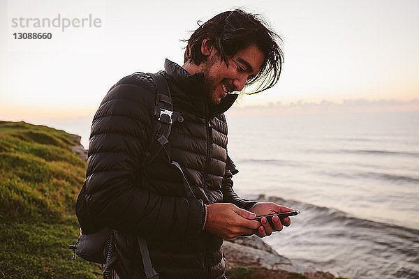 Glücklicher männlicher Wanderer  der ein Smartphone benutzt  während er auf einem Hügel am Meer steht