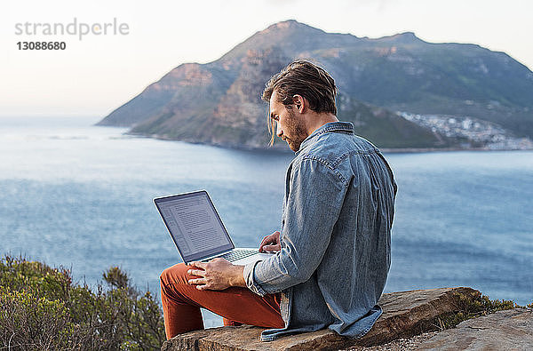 Seitenansicht eines Mannes mit Laptop auf einem Hügel gegen Berg und Meer