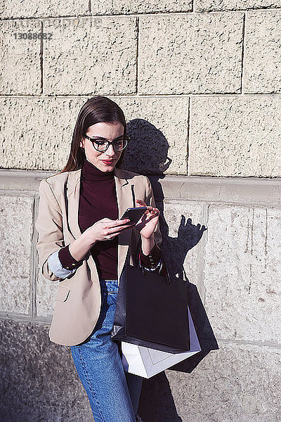 Frau mit Einkaufstaschen  die ein Mobiltelefon benutzt  während sie in der Stadt an einer Mauer steht
