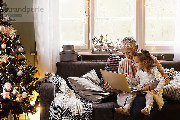 Glückliche Großmutter und Enkelin sitzen zu Hause auf dem Sofa und benutzen einen Laptop