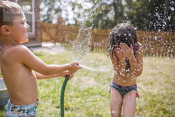 Bruder ohne Hemd bespritzt Schwester mit Wasser durch Gartenschlauch im Hof