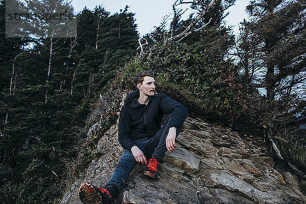 Nachdenklicher männlicher Wanderer sitzt auf Fels gegen Bäume