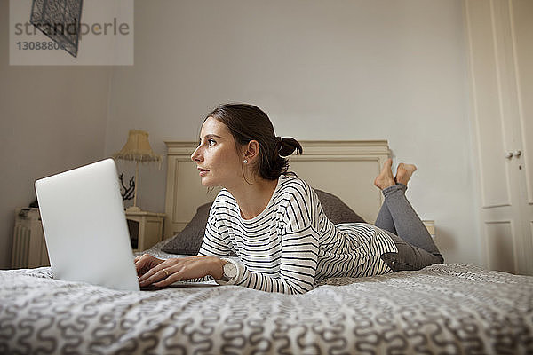 Nachdenkliche Frau mit Laptop  die zu Hause auf dem Bett liegt