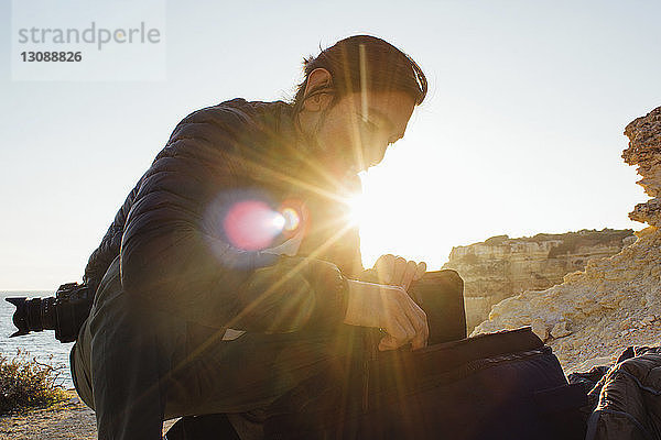 Niedrigwinkelansicht eines Mannes mit Rucksack  der am Strand vor klarem Himmel bei Sonnenuntergang kauert