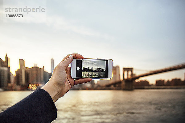 Geschnittene Hand einer Frau  die in der Dämmerung die Skyline und die Brooklyn Bridge fotografiert