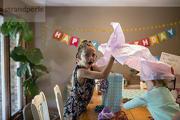 Porträt eines überraschten Mädchens mit Schwester beim Öffnen von Geburtstagsgeschenken auf dem Tisch zu Hause