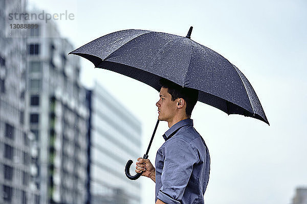 Seitenansicht eines jungen Geschäftsmannes mit Regenschirm  der bei Regen gegen Gebäude und Himmel in der Stadt steht