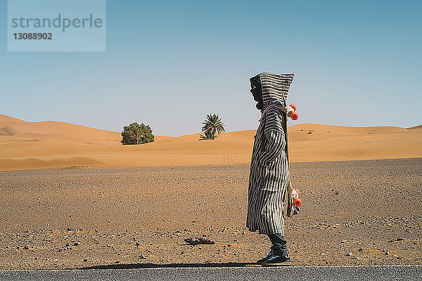 Seitenansicht eines Mannes mit Skateboard auf einer Straße in der Wüste gegen den Himmel bei Sonnenschein
