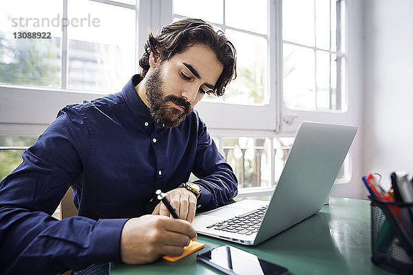 Geschäftsmann schreibt auf Haftnotiz  während er mit seinem Laptop im Büro am Tisch sitzt