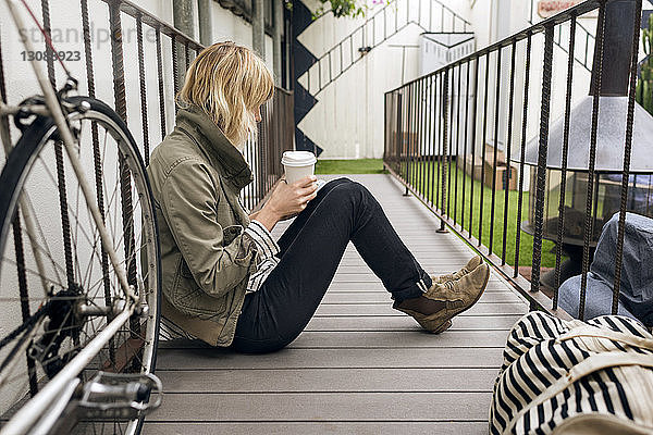 Seitenansicht einer jungen Frau  die Einwegglas hält  während sie auf einem Fußweg im Freien sitzt