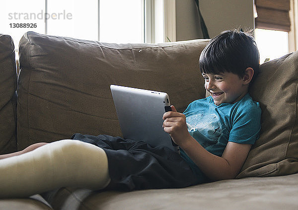 Lächelnder Junge mit gebrochenem Bein  der einen Tablet-Computer benutzt  während er zu Hause auf dem Sofa liegt