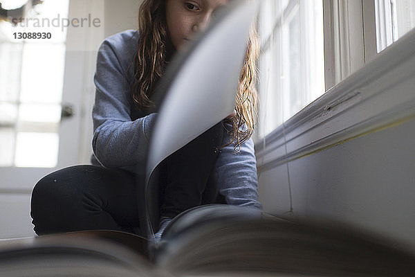 Mädchen liest Buch  während sie zu Hause am Fenster sitzt