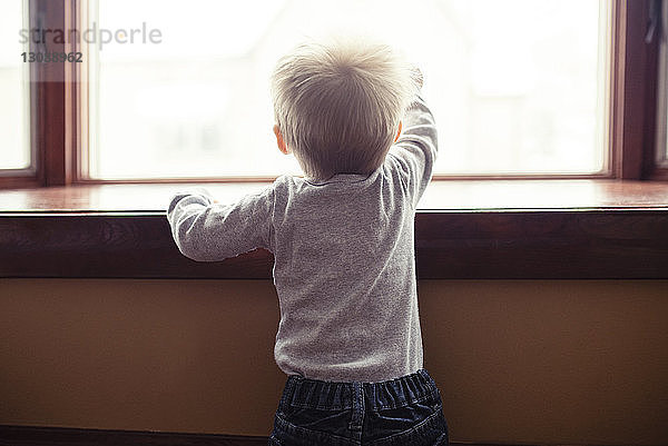 Rückansicht eines kleinen Jungen  der zu Hause durch ein Fenster schaut