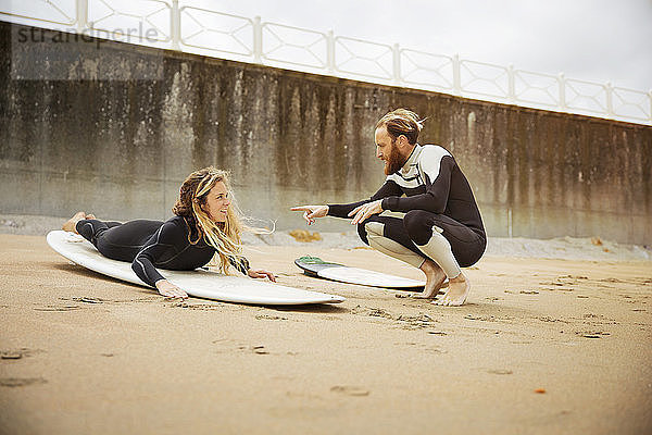 Mann spricht mit Frau  die am Strand auf einem Surfbrett liegt