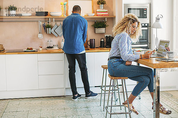 Frau benutzt Smartphone  während der Mann an der Küchentheke arbeitet