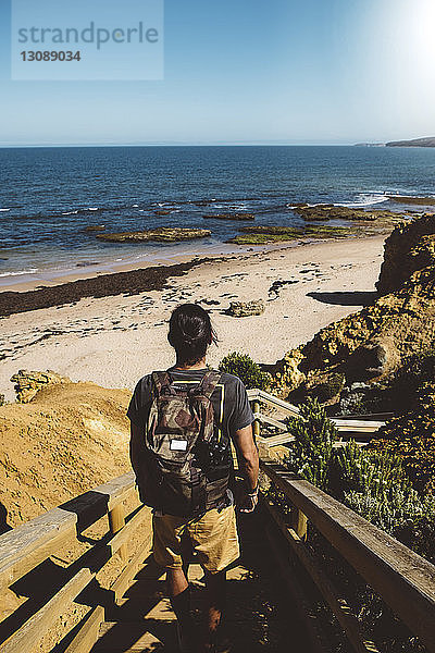 Rückansicht eines Mannes mit Rucksack  der auf Stufen am Strand vor strahlend blauem Himmel am sonnigen Tag steht