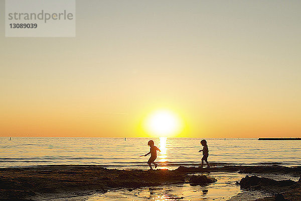 Scherenschnitt-Geschwister laufen am Strand gegen den klaren Himmel bei Sonnenuntergang