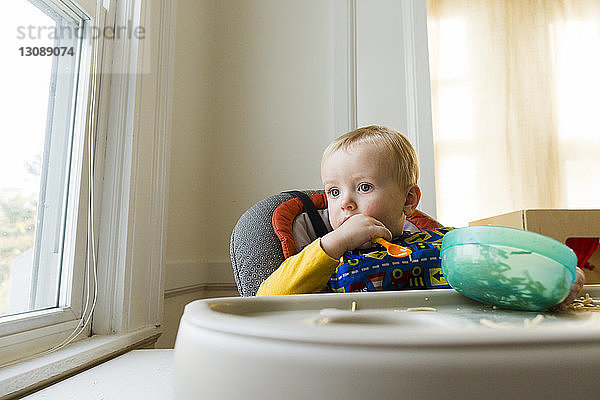 Süßer nachdenklicher kleiner Junge mit Plastikschüssel und Löffel  der zu Hause auf einem Hochstuhl sitzt
