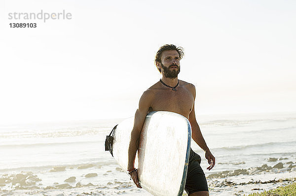Mann ohne Hemd mit Surfbrett beim Strandspaziergang vor klarem Himmel