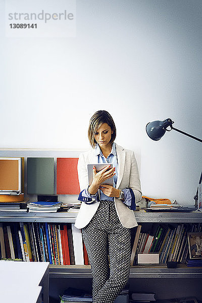 Geschäftsfrau benutzt Tablet-Computer  während sie sich im Büro auf einem Tisch an die Wand lehnt