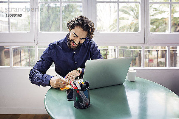 Glücklicher Geschäftsmann schreibt auf Klebezettel  während er mit seinem Laptop im Büro am Tisch sitzt
