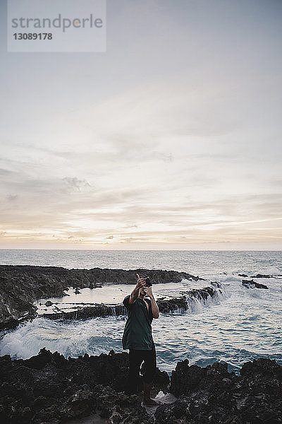 Mann zeigt obszöne Geste  während er auf Felsen am Meer steht