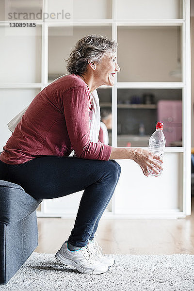 Seitenansicht einer sportlich reifen Frau  die eine Wasserflasche hält  während sie zu Hause auf dem Sofa sitzt