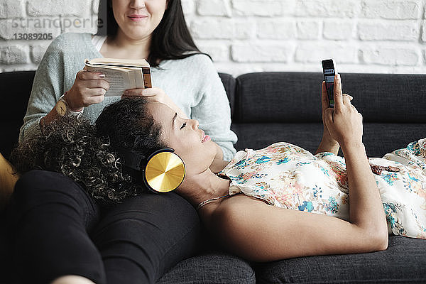Frau benutzt Mobiltelefon  während sie sich auf dem Schoß ihrer Freundin entspannt und im Wohnzimmer ein Buch liest